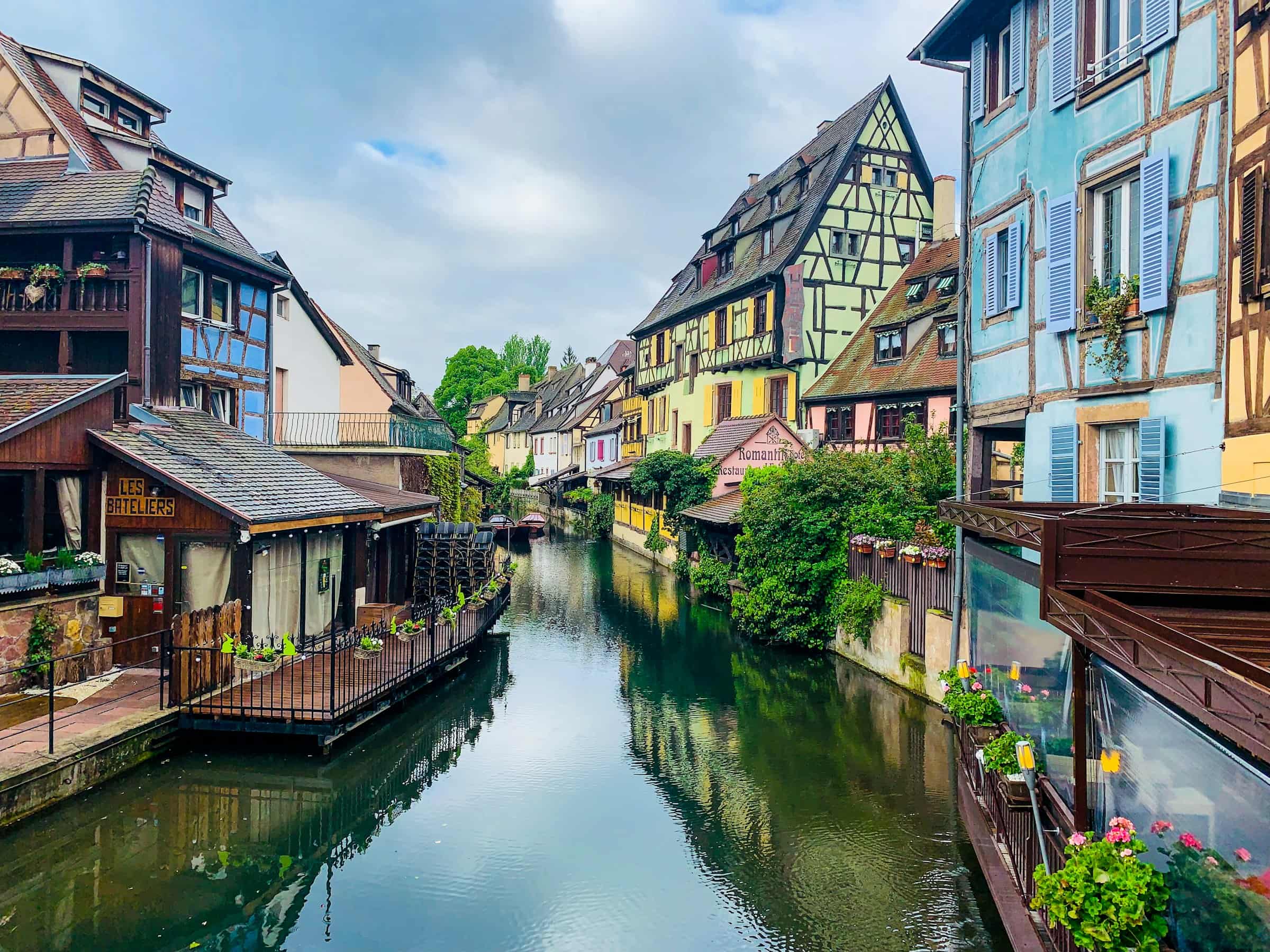 Un canal étroit vu depuis un pont, bordé sur ses deux rives de maisons à colombages — une brune à balcon fleuri et son embarcadère à gauche, une bleu pâle et une vert d’eau à droite —, des barques amarrées, des géraniums aux balustrades et un ciel gris de nuages