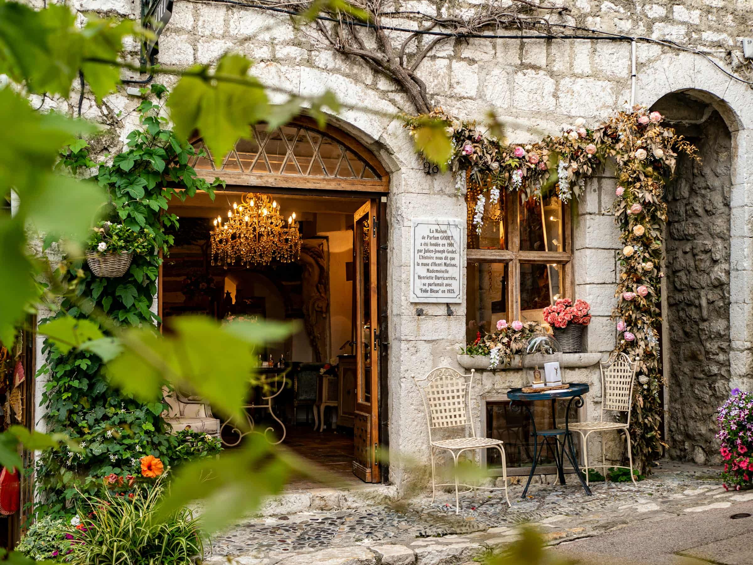 Une maison de village en pierre claire, sa façade couverte de fleurs, volets ouverts, en plein jour