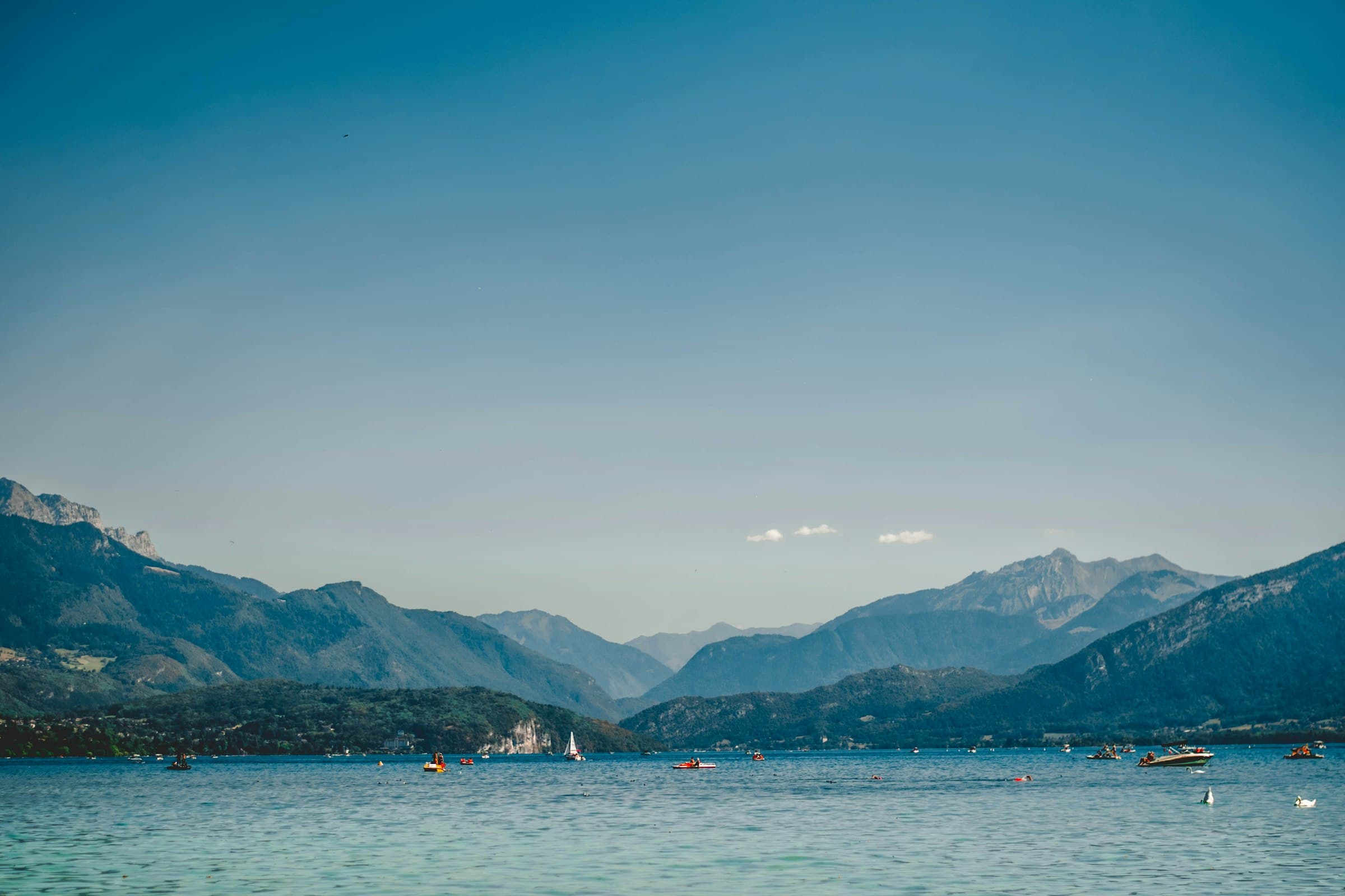 Le lac d’Annecy depuis la rive, l’eau turquoise au premier plan, quelques barques au loin et la ligne des montagnes sur l’autre rive