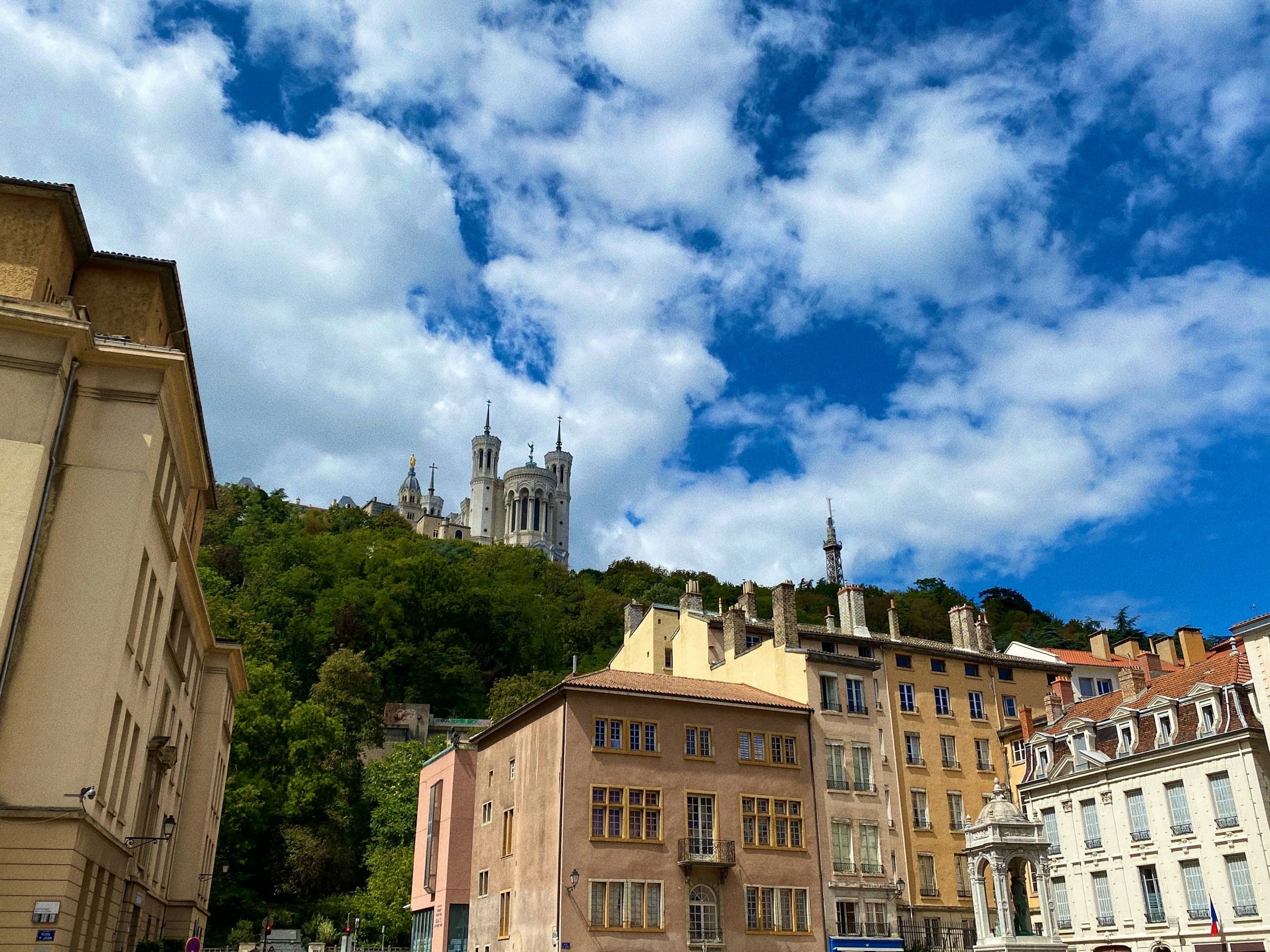 Une basilique blanche dressée au sommet d’une colline, au-dessus de façades ocre, sous un ciel bleu chargé de nuages blancs