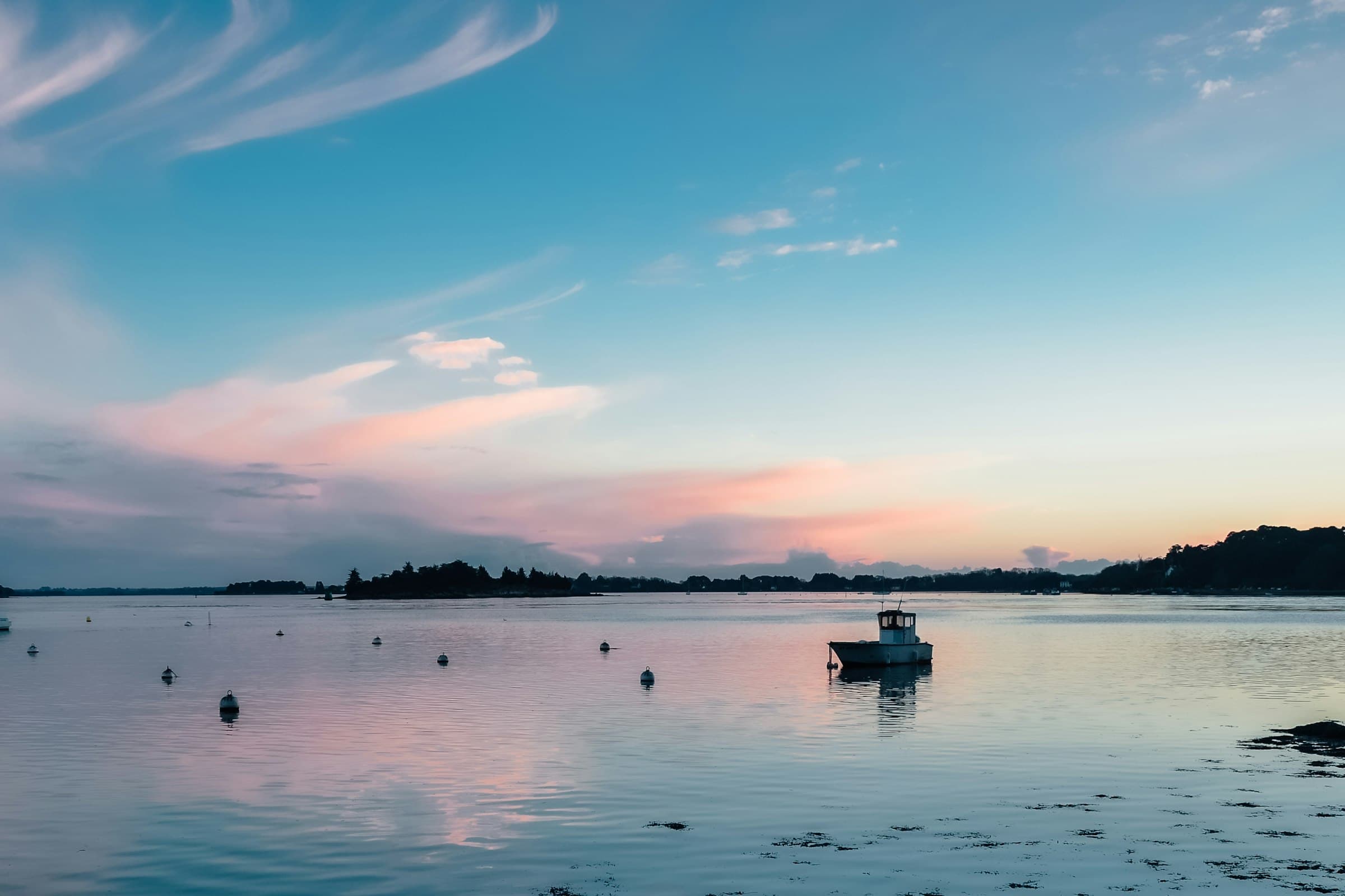 Un petit bateau de pêche au mouillage sur une eau étale au crépuscule, un chapelet de bouées devant lui, la rive boisée réduite à une silhouette et un ciel de cirrus roses et bleus