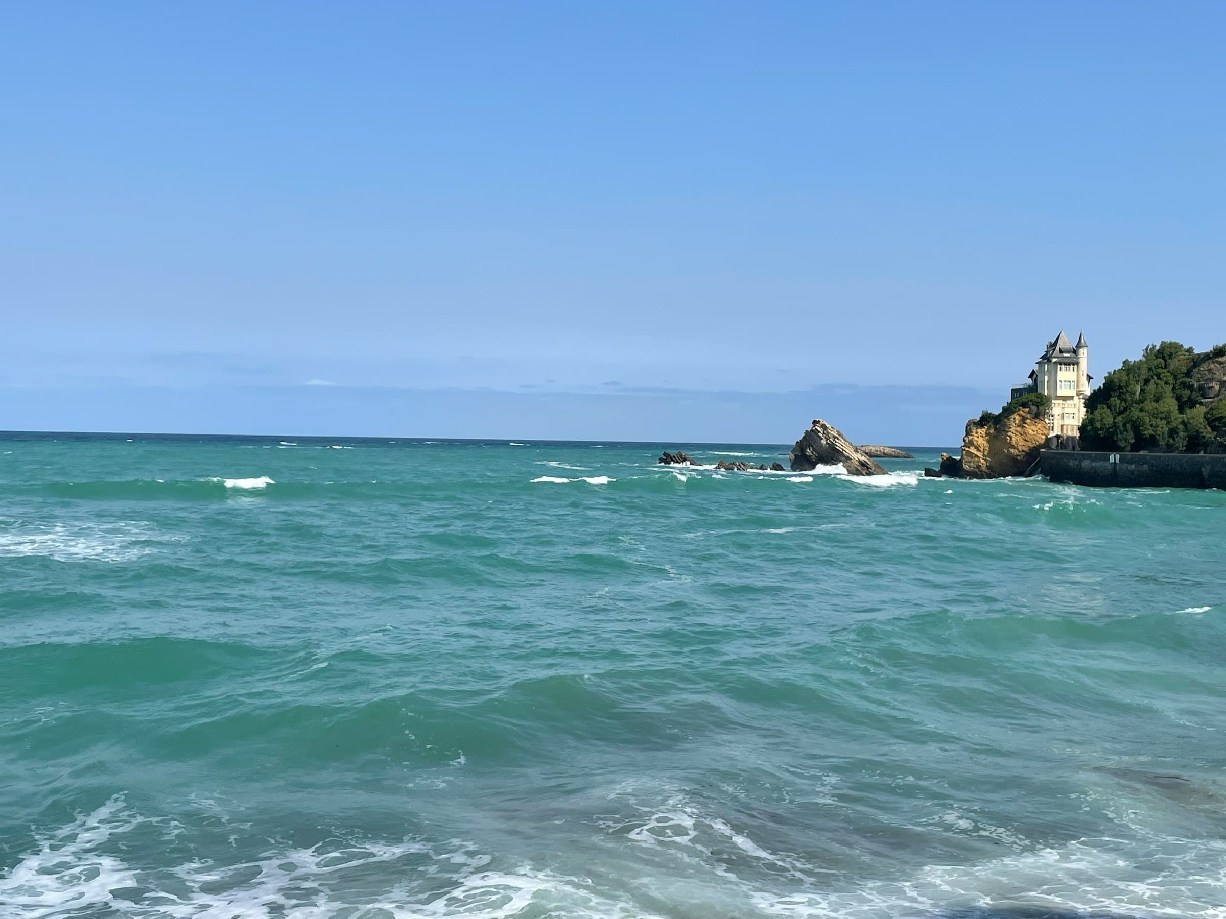 La mer verte et l’écume devant la villa Belza de Biarritz, sa tourelle posée sur le rocher à droite, sous un ciel bleu franc
