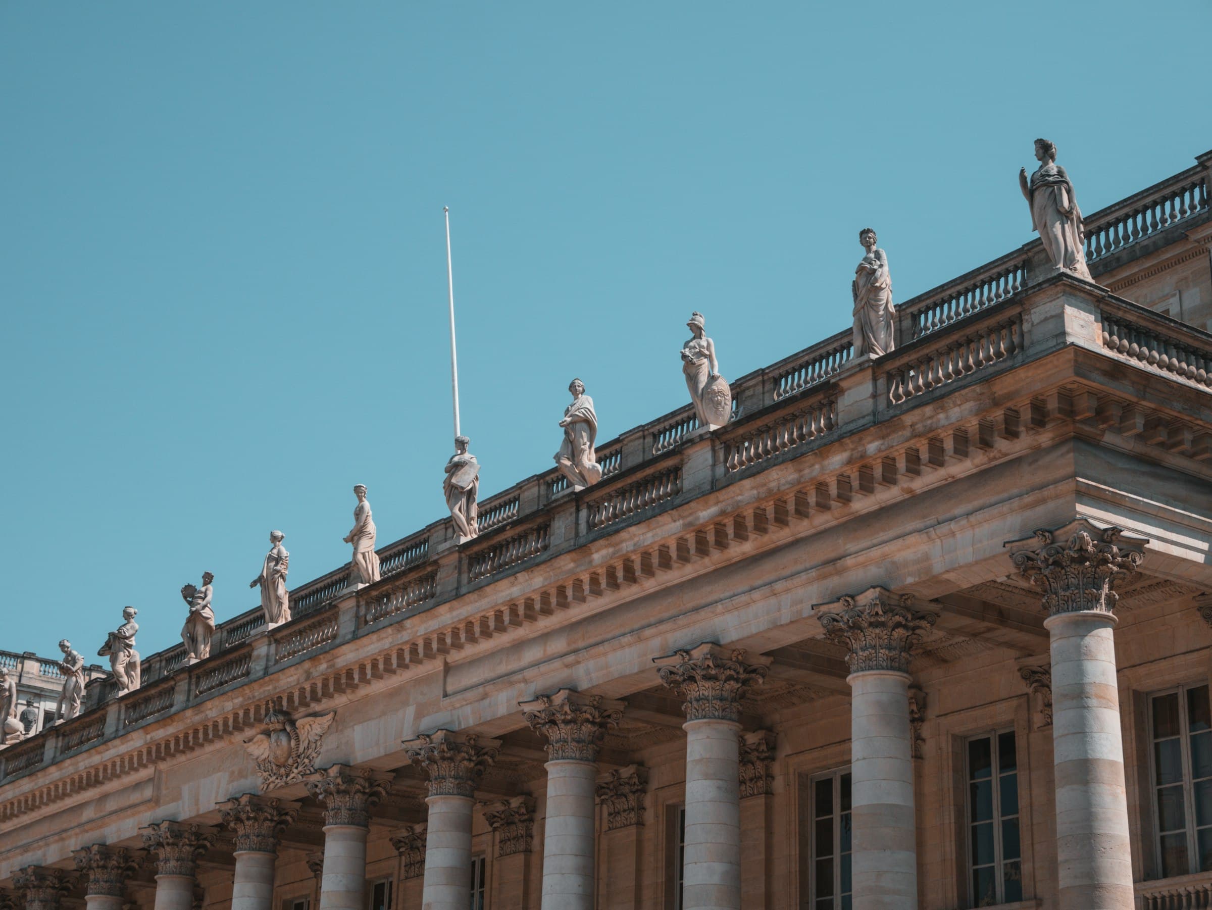 La colonnade du Grand Théâtre de Bordeaux vue en contre-plongée, les statues alignées sur la corniche et le ciel bleu profond derrière