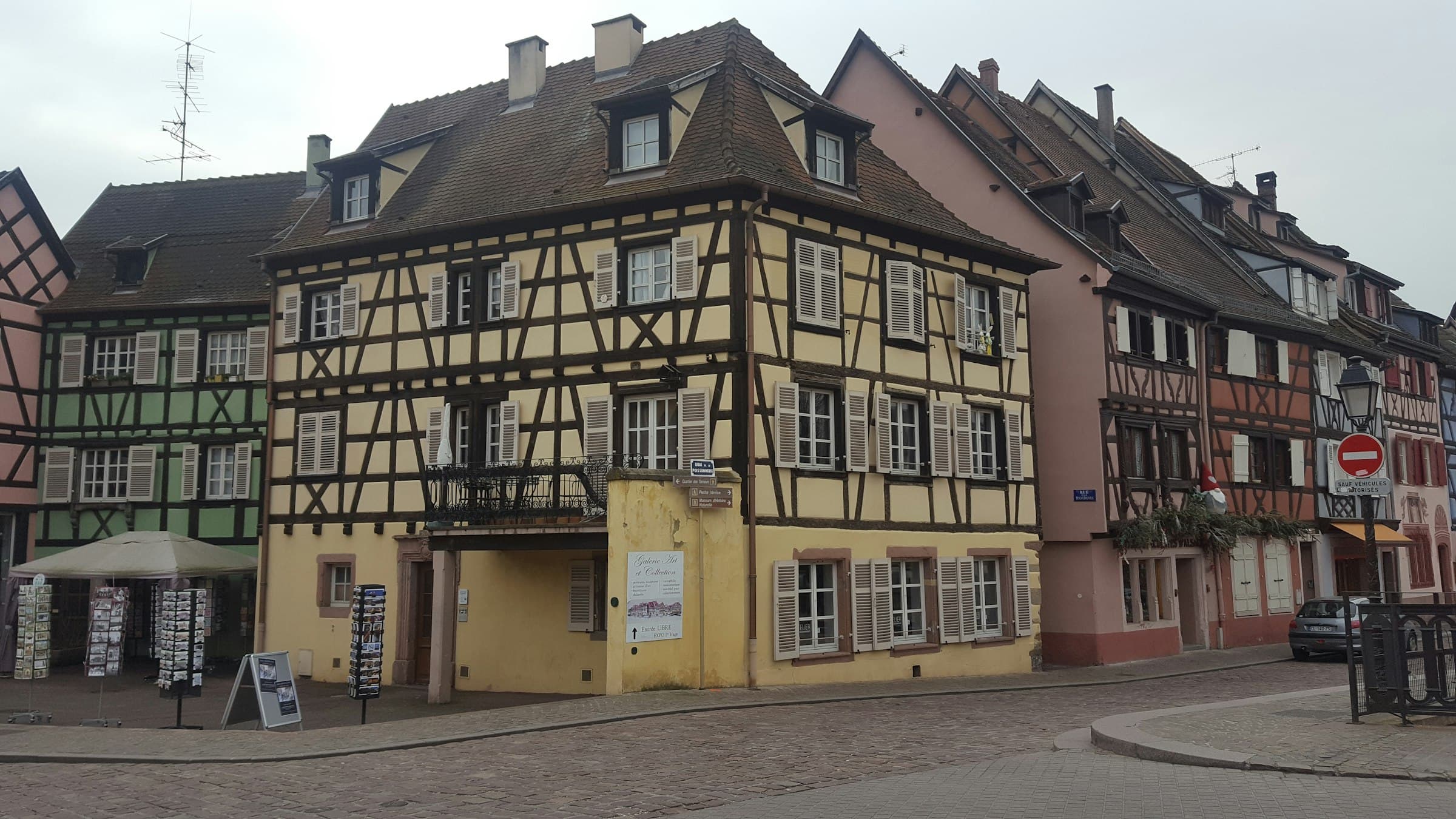 Un angle de rue pavée du vieux Colmar : une maison à colombages jaune à pans de bois bruns, ses voisines vert d’eau et rose, volets gris clair et présentoir de cartes postales, sous un ciel couvert