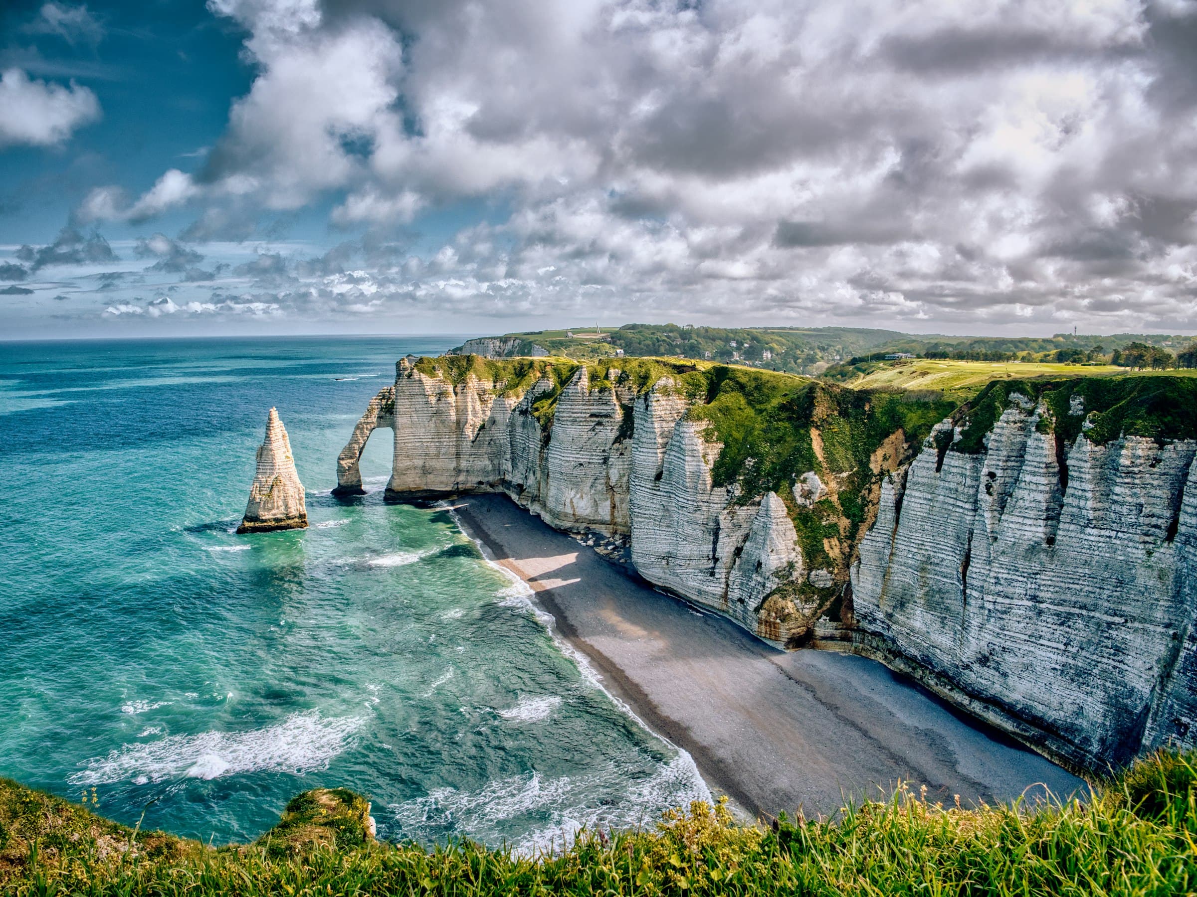 La falaise d’Aval et l’aiguille d’Étretat vues depuis les herbes du plateau, la mer turquoise et la plage de galets en contrebas, sous un ciel de nuages