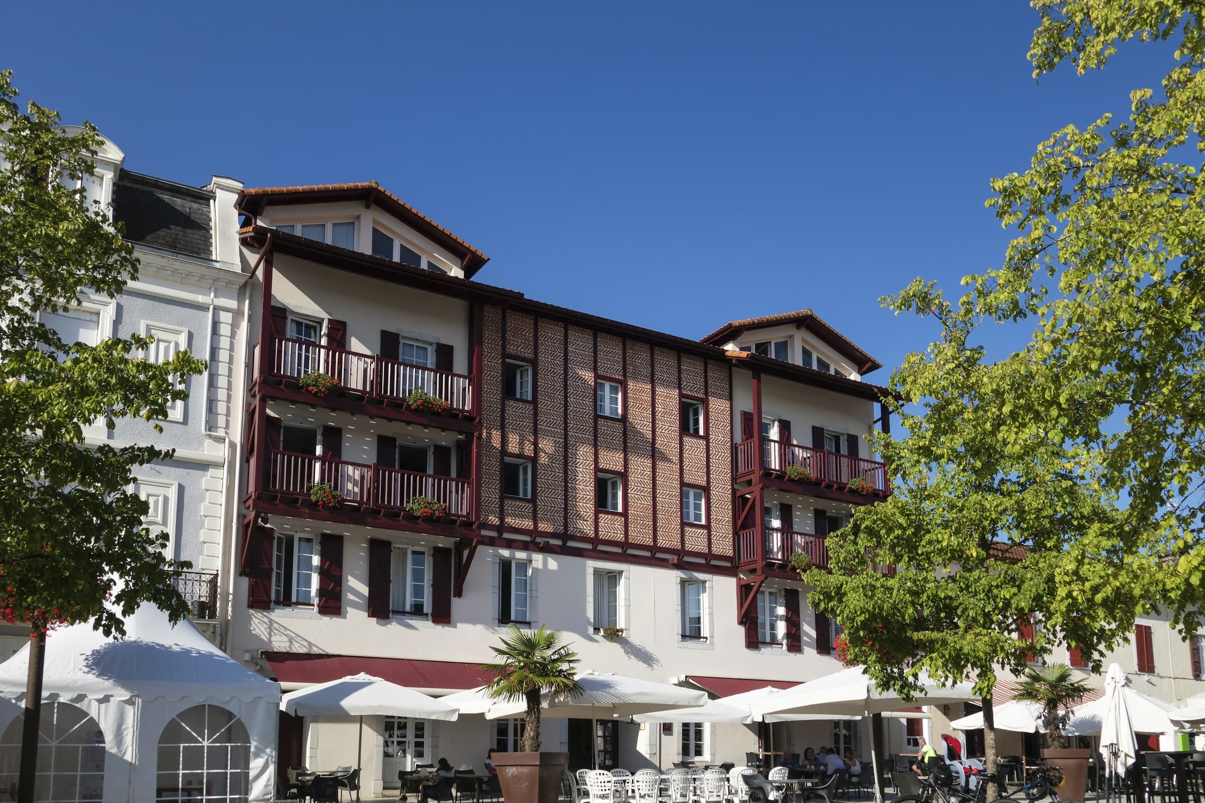 Une maison traditionnelle du Pays basque, colombages rouges sur enduit blanc et balcons de bois, au-dessus des parasols d’une terrasse