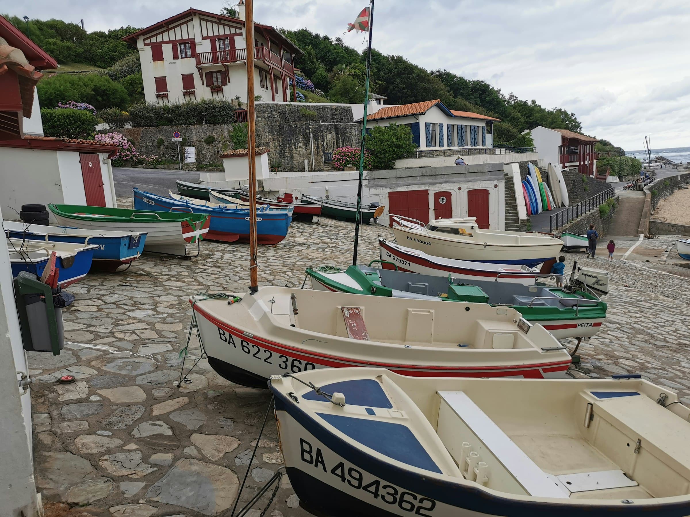 Des barques de pêche tirées sur la cale pavée d’un petit port de la côte basque, des maisons blanches aux volets rouges derrière