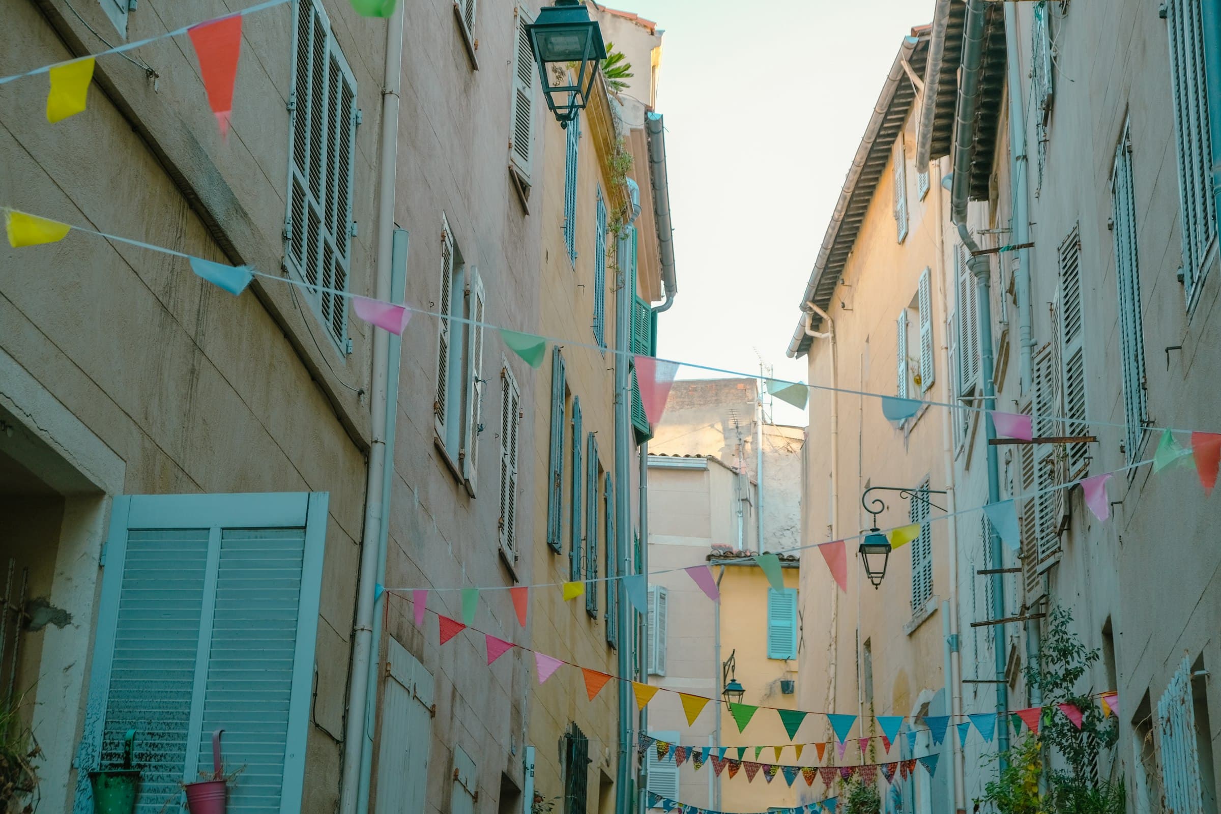 Une rue étroite bordée de façades pastel aux volets bleus, des fanions colorés tendus d’un immeuble à l’autre au-dessus de la chaussée