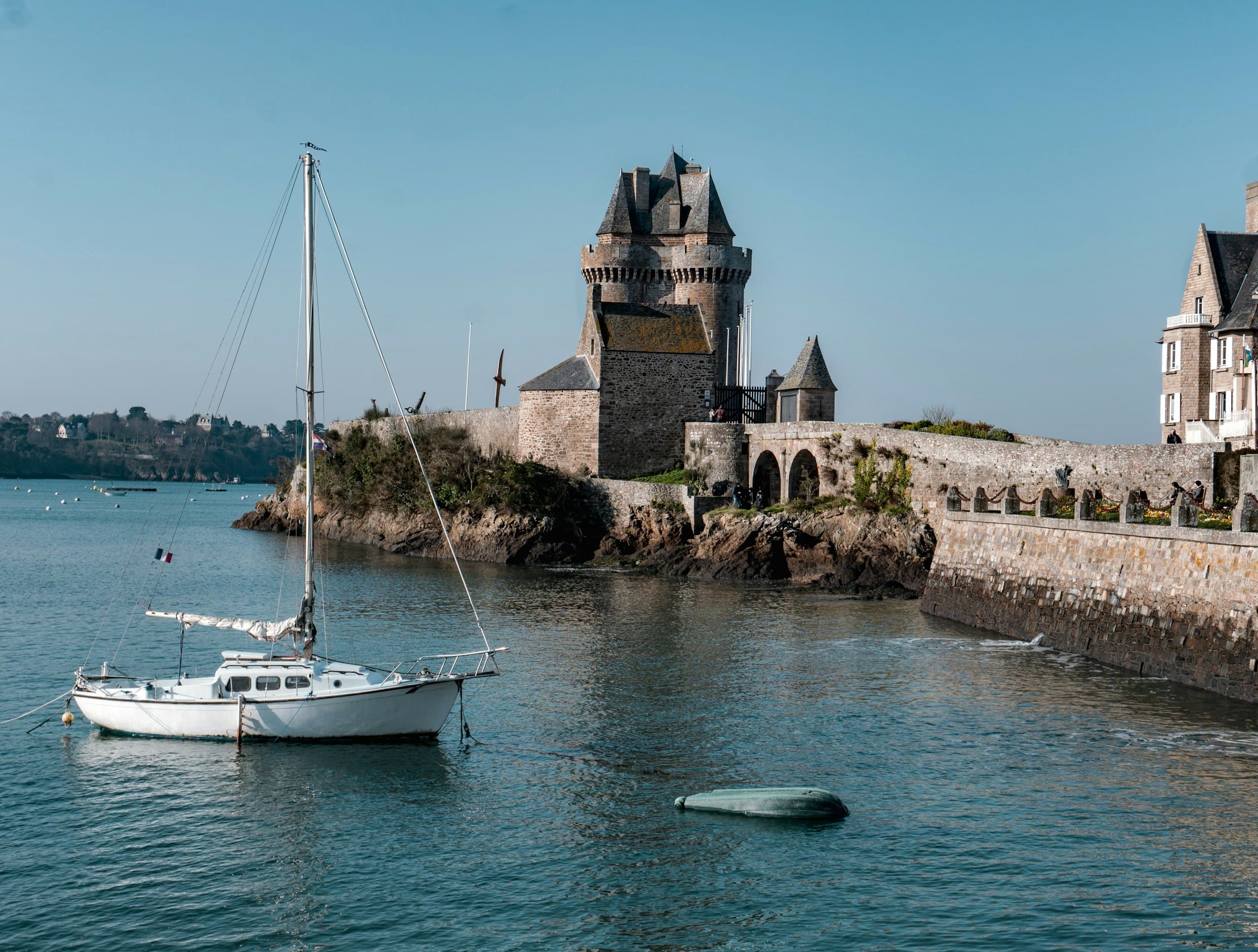 Un voilier blanc au mouillage devant la tour de granit à trois tourelles qui garde l’embouchure, à Saint-Malo, le promontoire rocheux et une digue de pierre bordée de maisons à droite, sous un ciel bleu clair