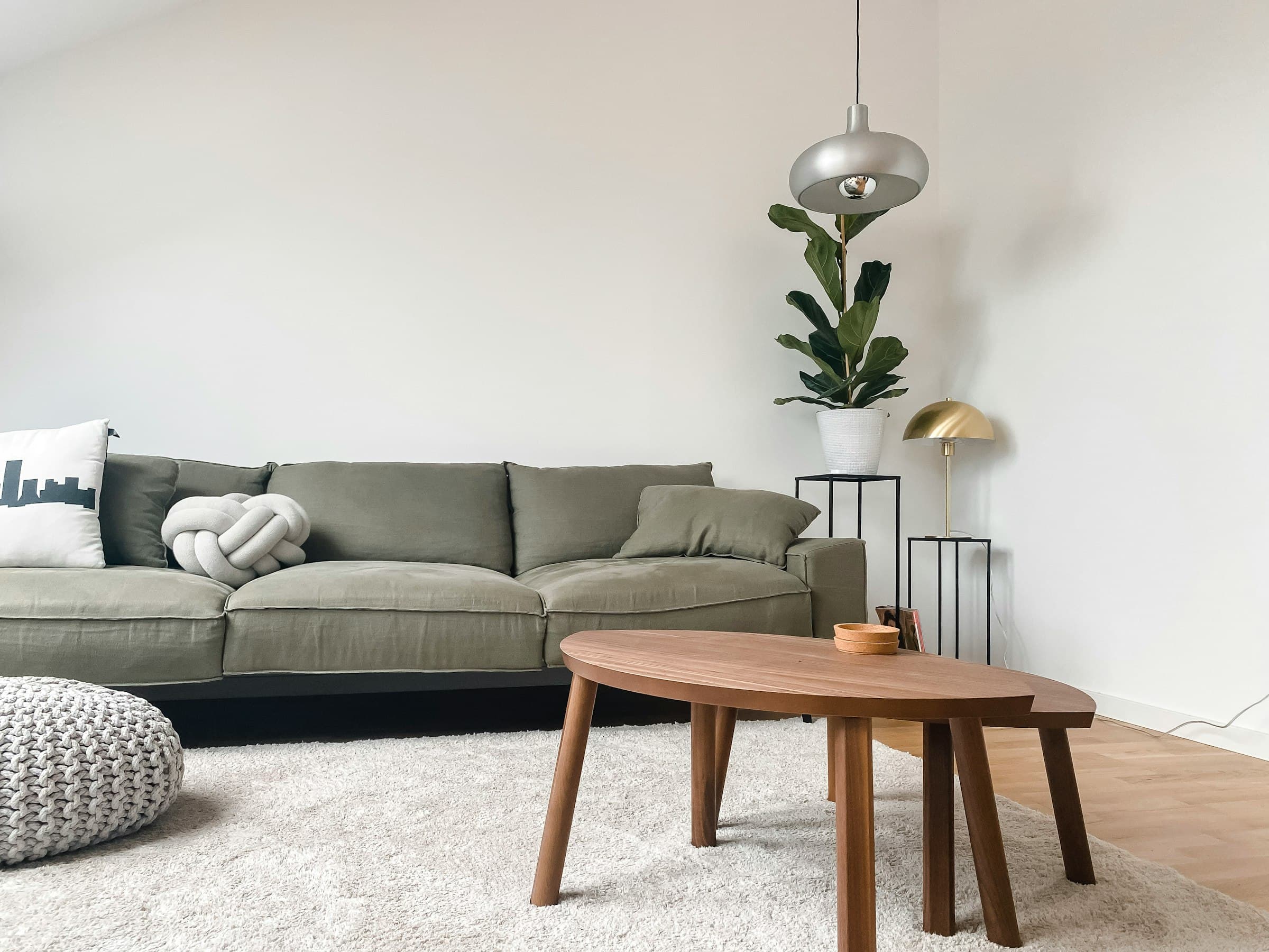 Un salon clair et rangé : un canapé de lin vert-de-gris, une table basse ovale en bois, un tapis épais, un ficus lyre et deux lampes en laiton contre un mur blanc