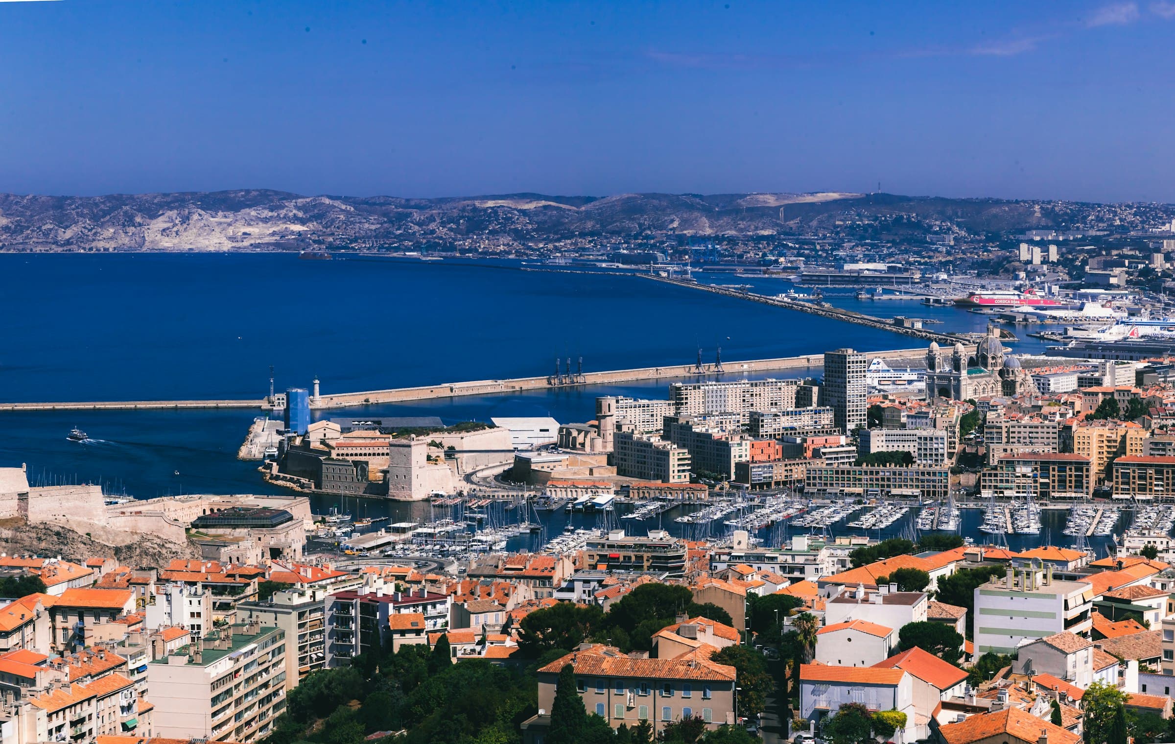 Une ville portuaire vue depuis les hauteurs : les toits de tuiles au premier plan, les bassins du port et la rade bleue jusqu’aux collines