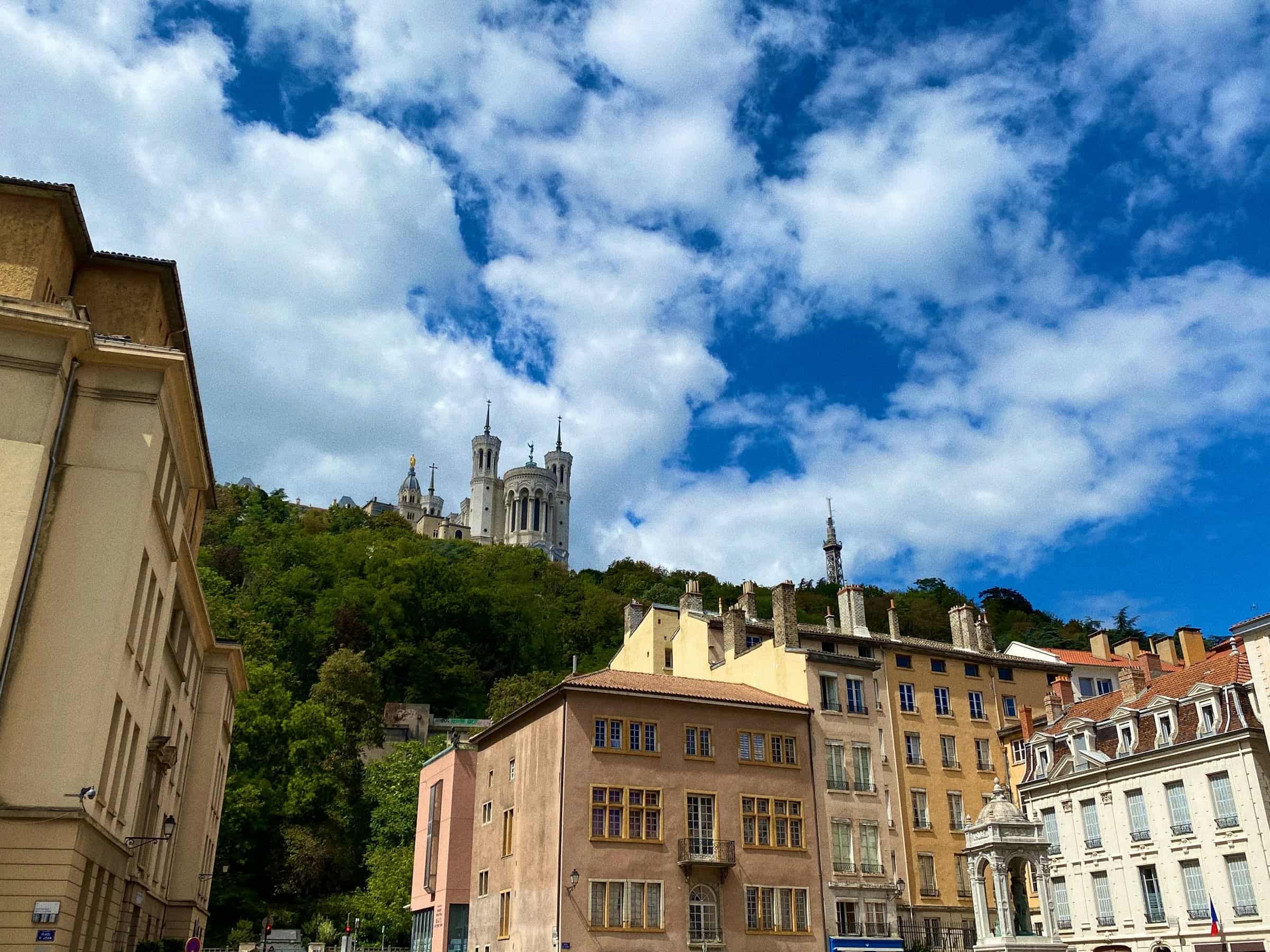 Une basilique blanche dressée au sommet d’une colline, au-dessus de façades ocre, sous un ciel bleu chargé de nuages blancs