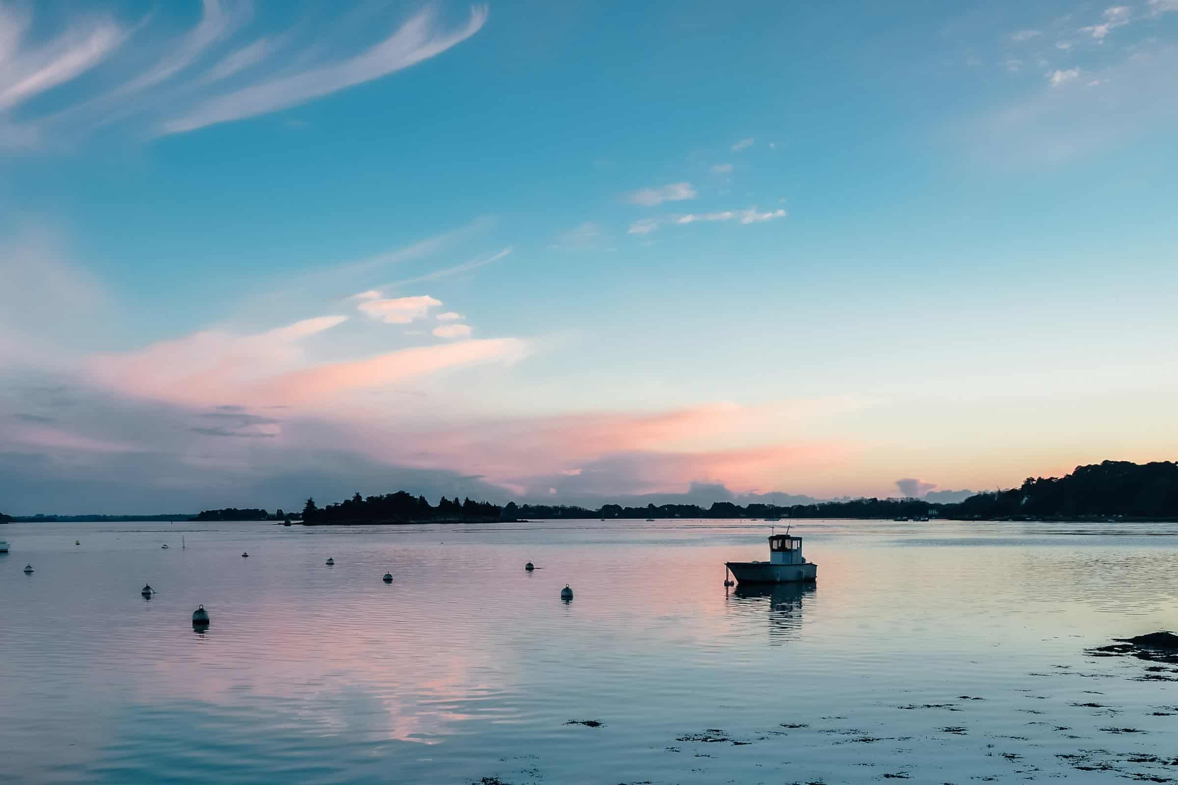 Un petit bateau de pêche au mouillage sur une eau étale au crépuscule, un chapelet de bouées devant lui, la rive boisée réduite à une silhouette et un ciel de cirrus roses et bleus