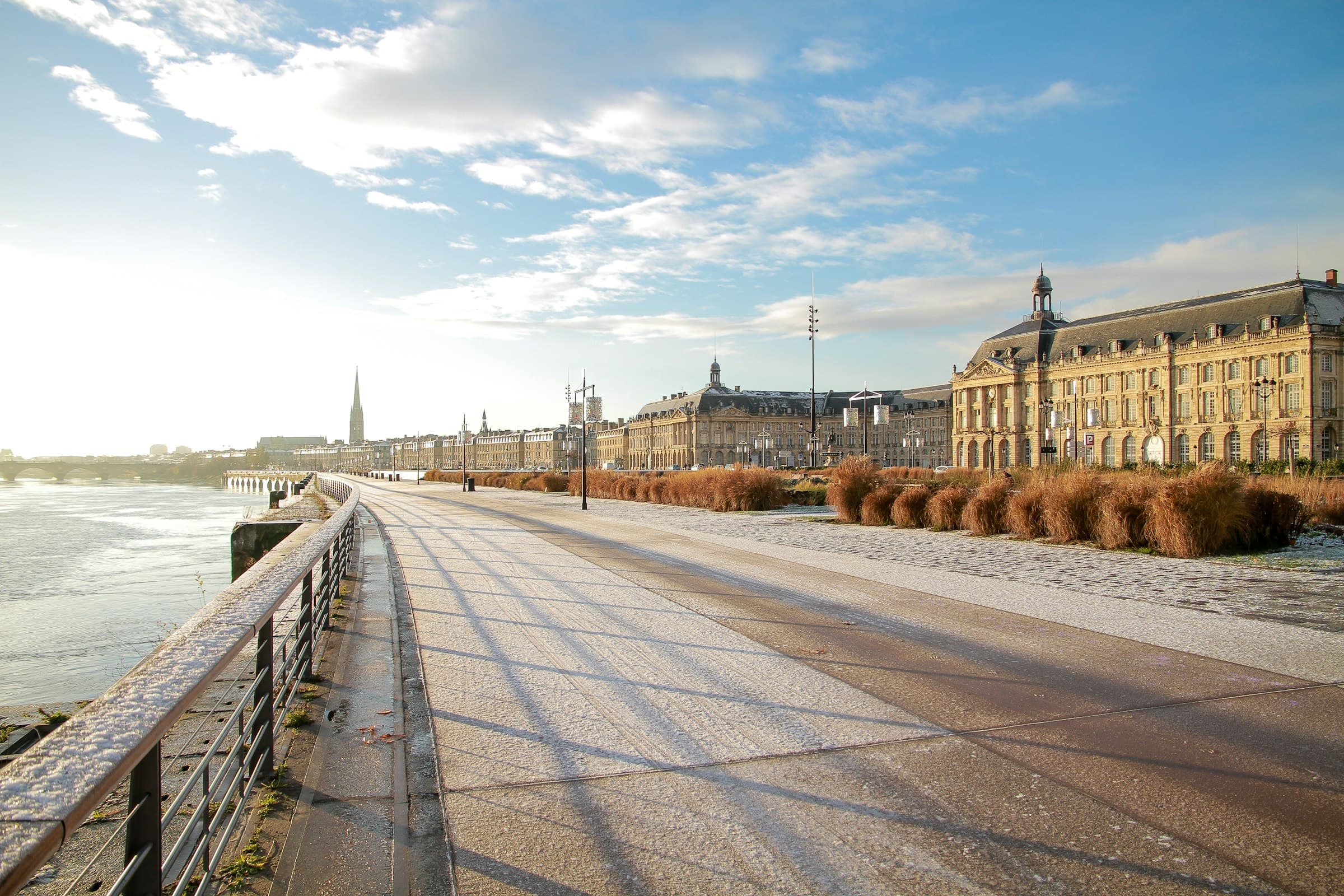 Les quais de Bordeaux un matin d’hiver : la Garonne à gauche, la promenade qui s’éloigne, les façades de pierre blonde et la place de la Bourse à droite sous un ciel bleu