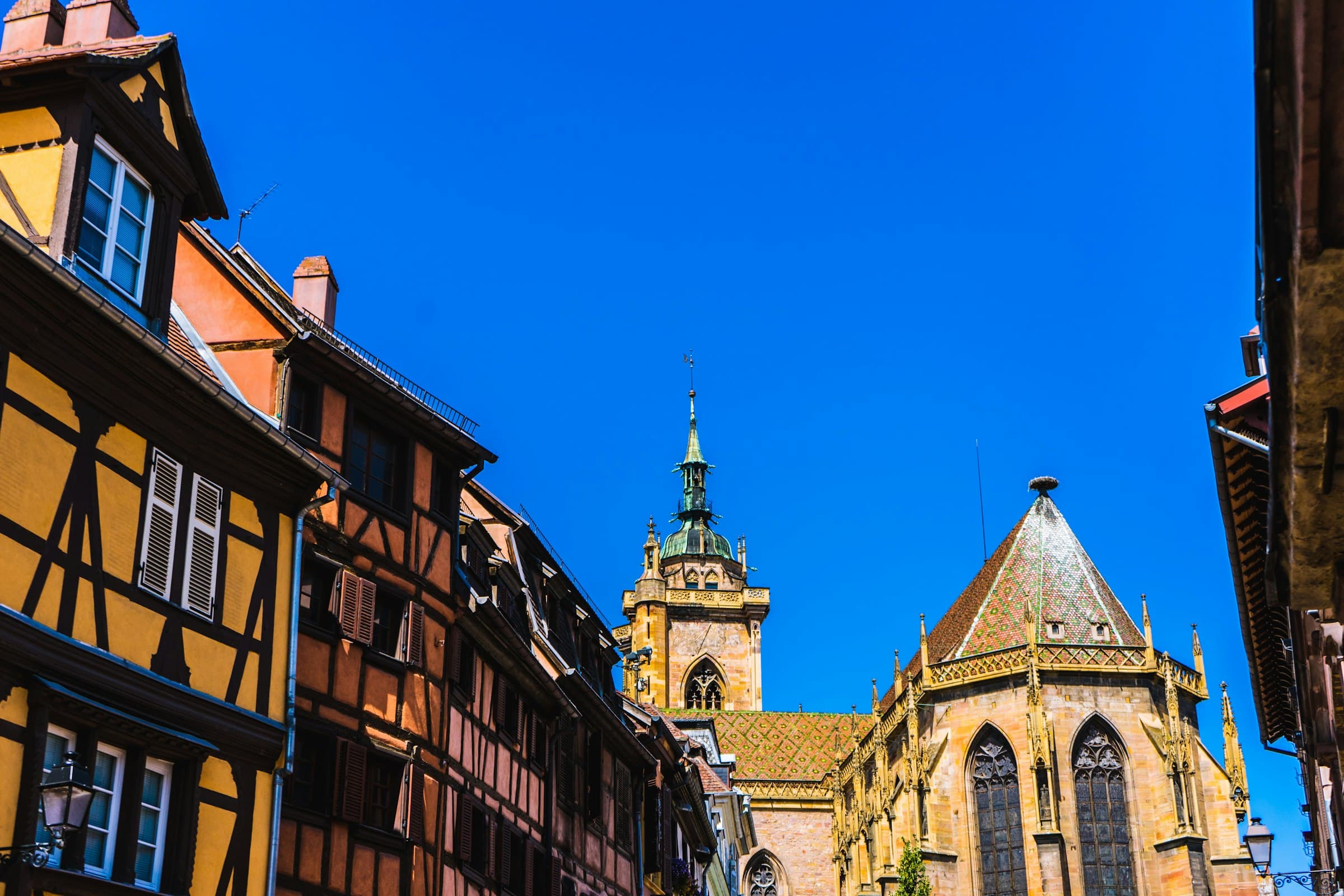 Une ruelle de bourg alsacien bordée de colombages jaunes et rouges, un clocher gothique se détachant sur un ciel bleu franc