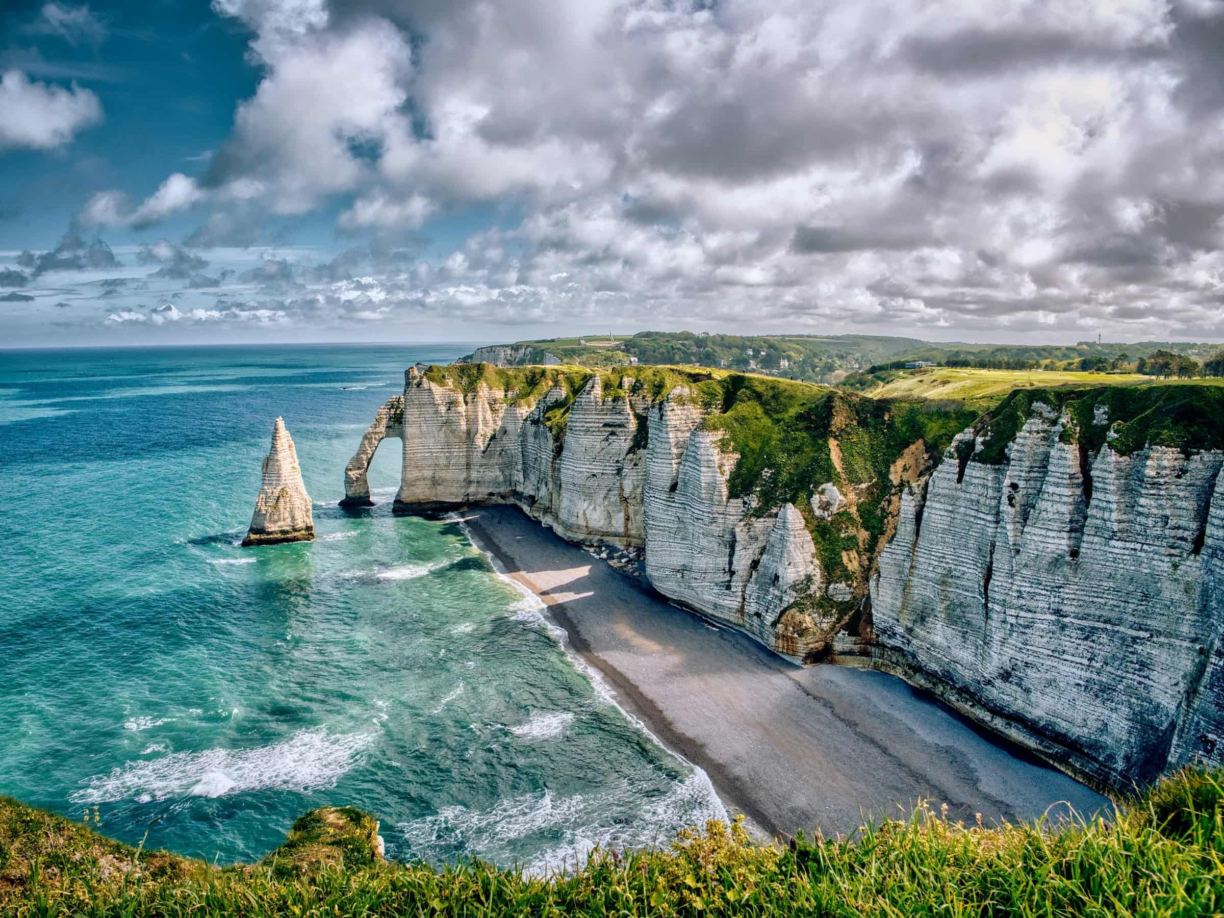 La falaise d’Aval et l’aiguille d’Étretat vues depuis les herbes du plateau, la mer turquoise et la plage de galets en contrebas, sous un ciel de nuages