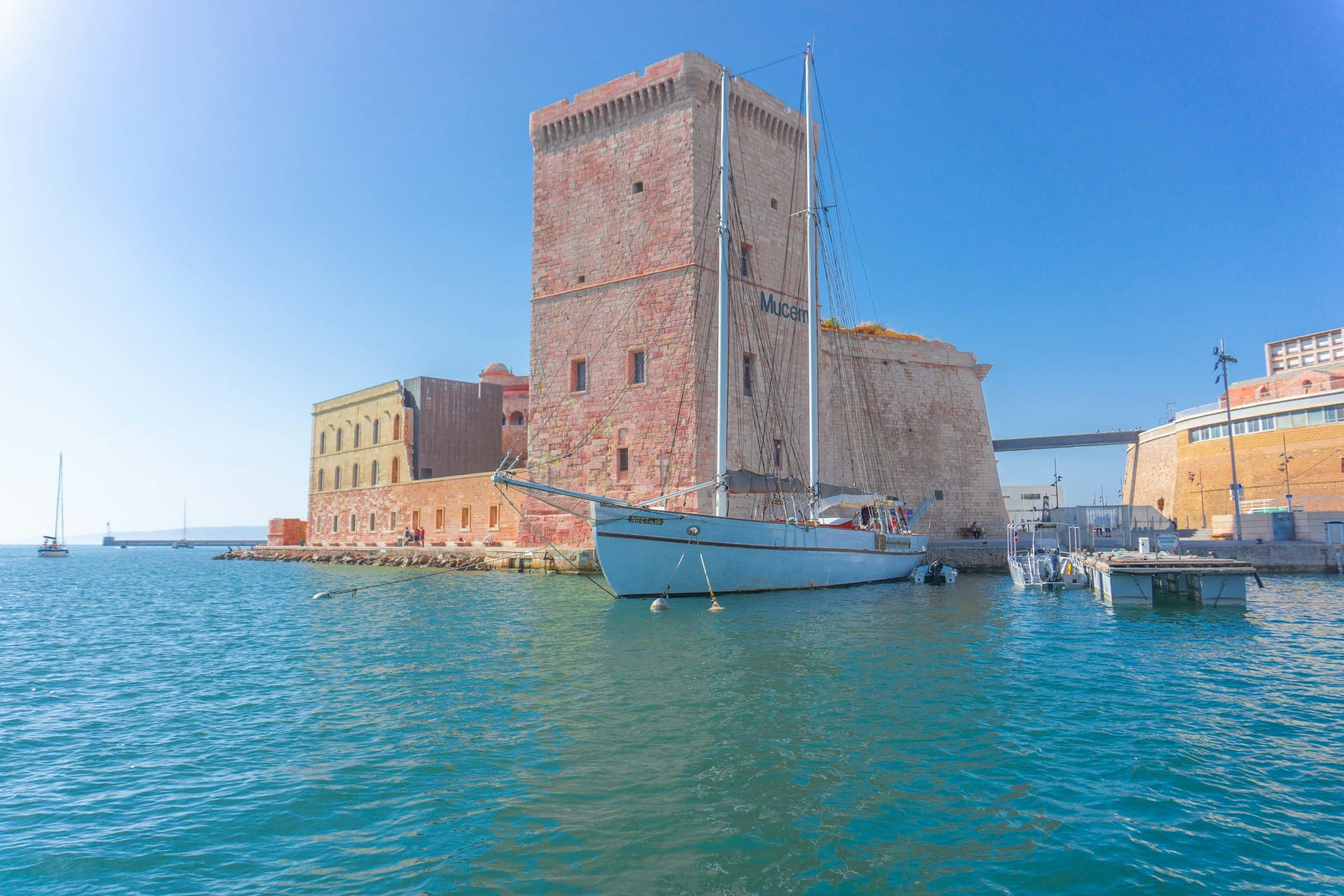 La tour de pierre du fort qui garde l’entrée du port de Marseille, la mer bleue au premier plan et une passerelle en arrière-plan sous un ciel dégagé