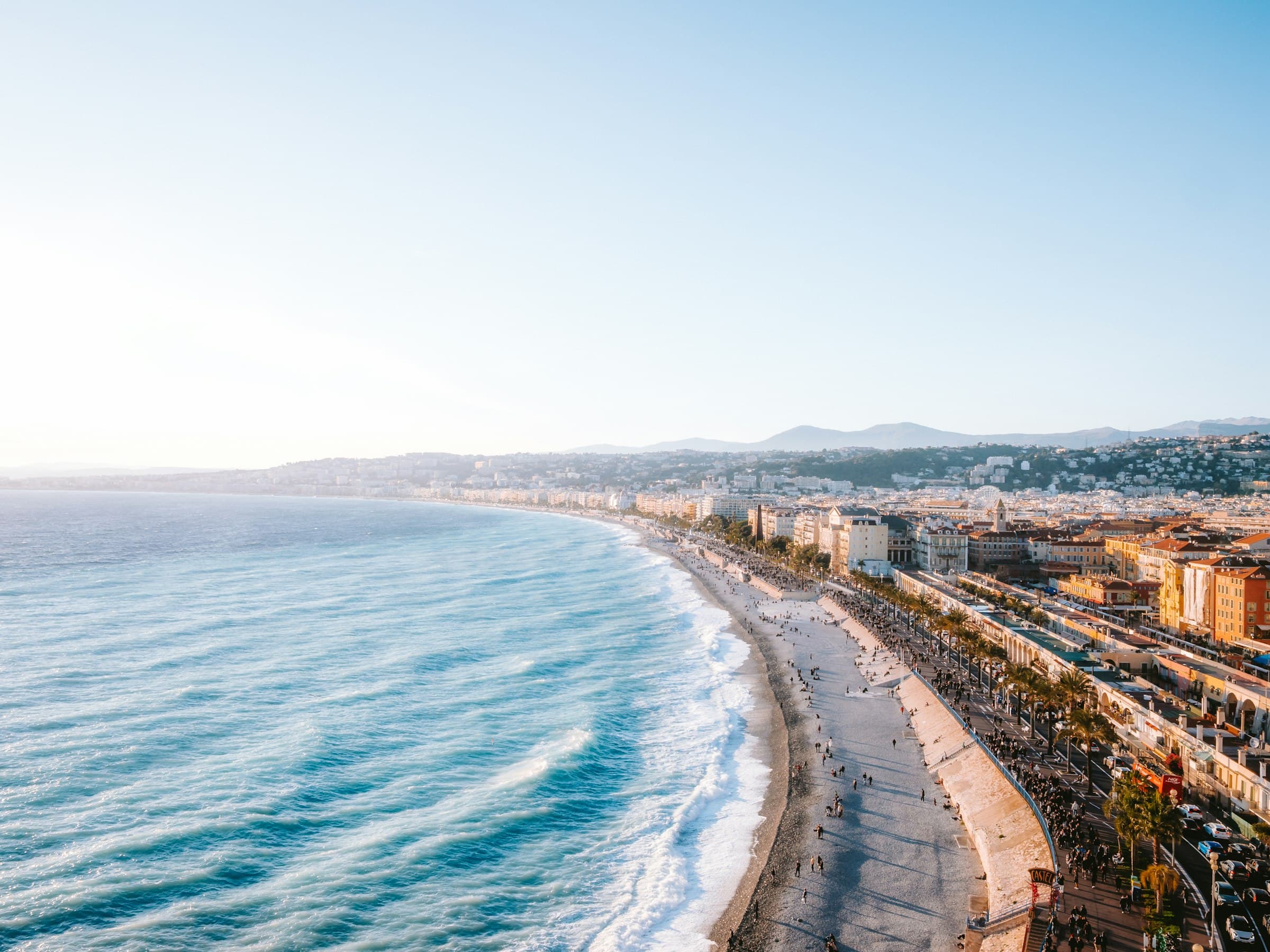 La courbe de la baie de Nice en plein jour, les vagues turquoise sur la plage de galets et la ville qui monte sur la colline derrière