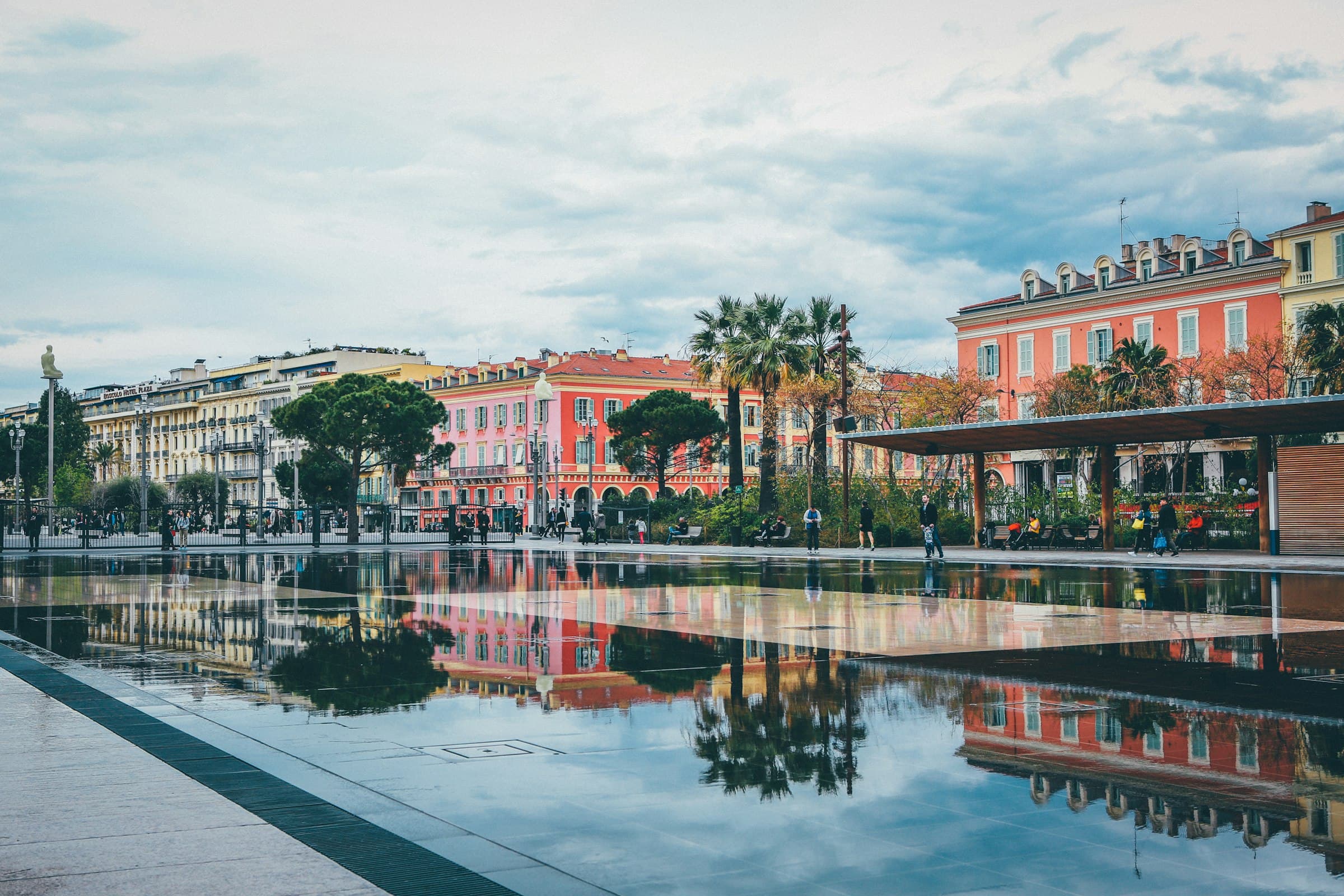 Un bassin miroir à Nice reflétant les façades ocre et les palmiers de la promenade, quelques passants au loin en plein jour