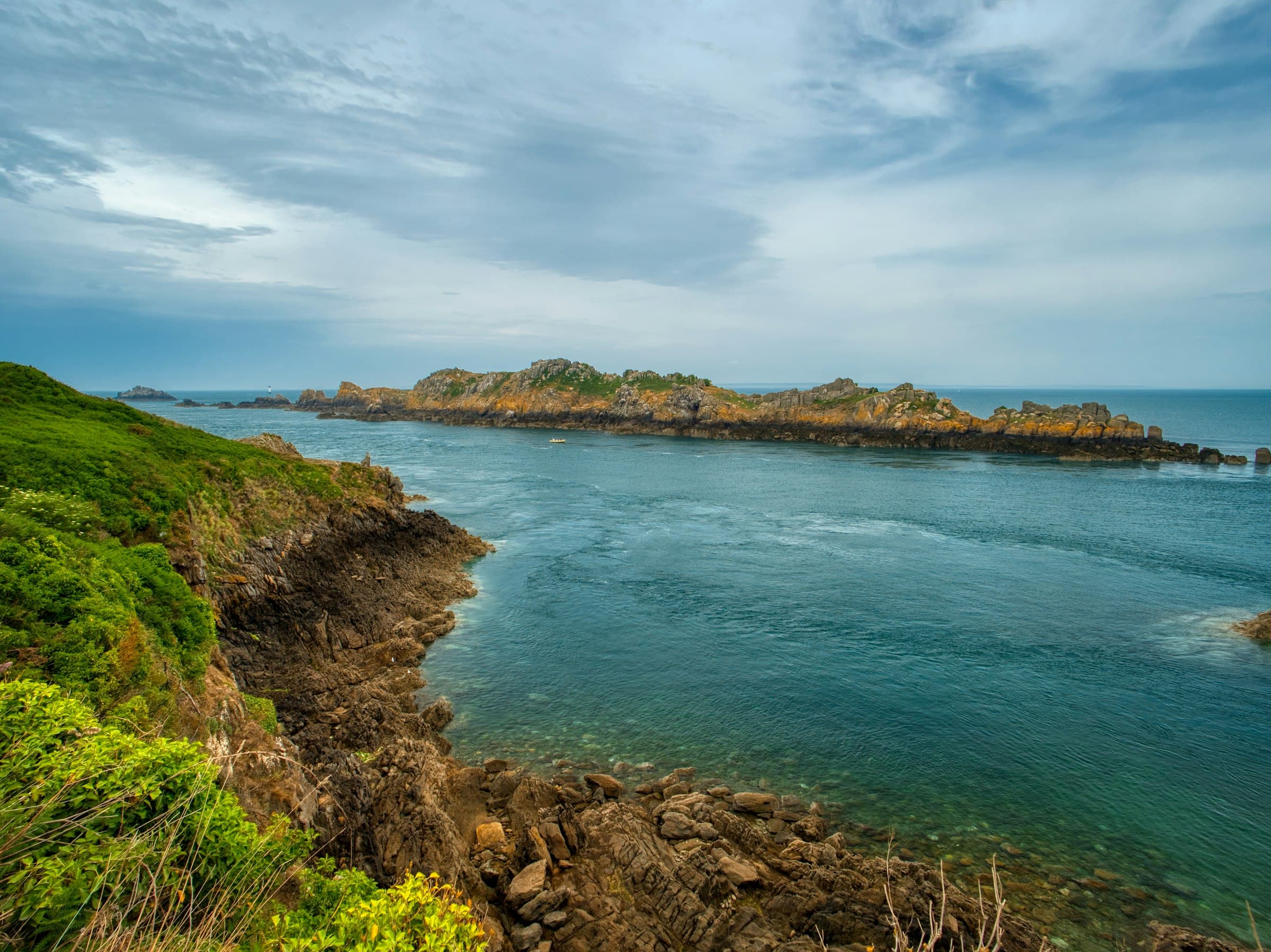 Une côte rocheuse en plein jour : au premier plan à gauche la lande, les ronces et la falaise de roche sombre et feuilletée ; au centre un bras de mer bleu-vert traversé de courants ; en face un long îlot bas hérissé de blocs clairs tachés de lichen orangé, une petite embarcation dans le passage, et à l’horizon un phare blanc sur son rocher, sous un ciel de gros nuages
