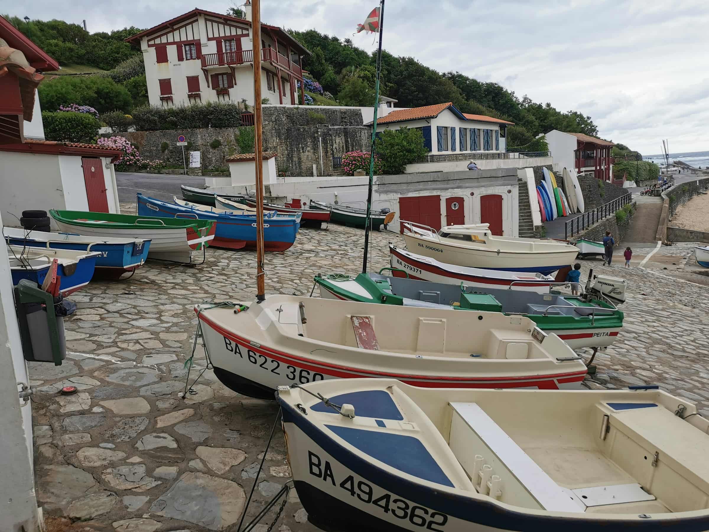 Des barques de pêche tirées sur la cale pavée d’un petit port de la côte basque, des maisons blanches aux volets rouges derrière