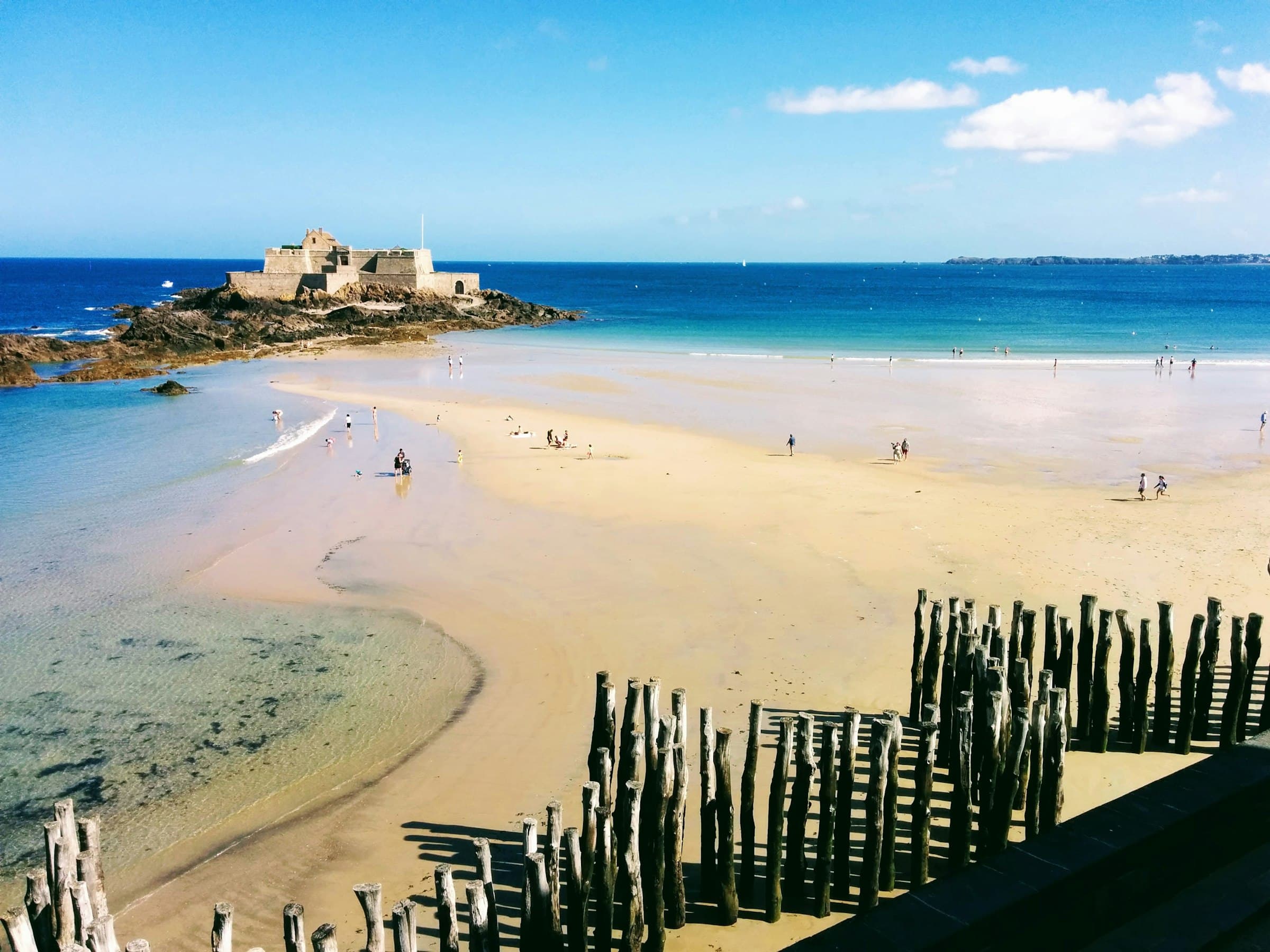 La grande plage de Saint-Malo à marée basse : les brise-lames de troncs plantés en quinconce au premier plan, le sable découvert sur toute la largeur, un fort sur son îlot rocheux à gauche et la mer turquoise à droite, sous un ciel de nuages blancs