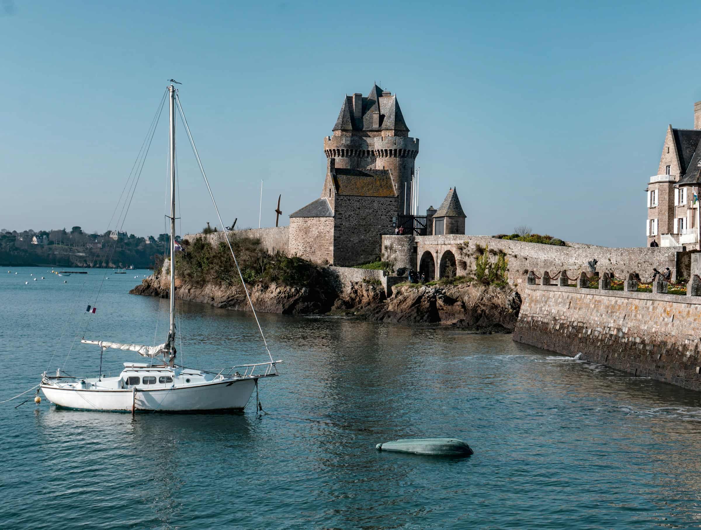 Un voilier blanc au mouillage devant la tour de granit à trois tourelles qui garde l’embouchure, à Saint-Malo, le promontoire rocheux et une digue de pierre bordée de maisons à droite, sous un ciel bleu clair