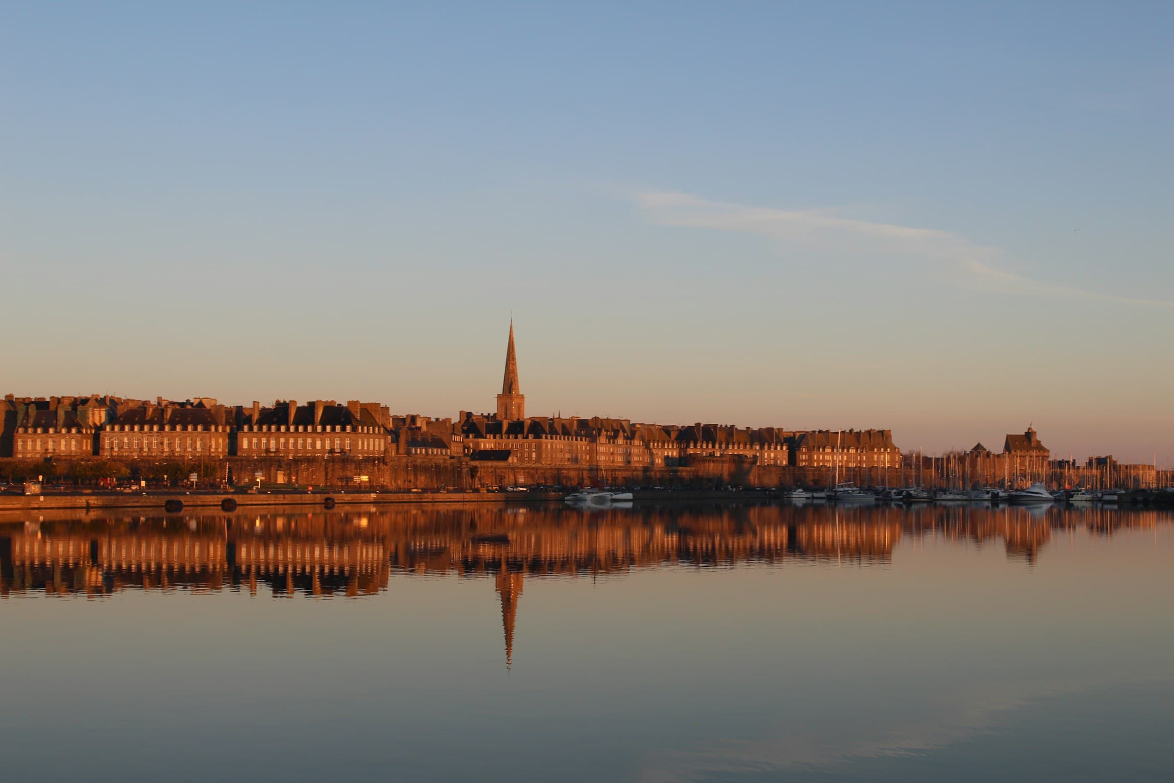 La ville close de Saint-Malo vue depuis le plan d’eau du port : le rempart de granit, l’alignement des hautes maisons, la flèche de la cathédrale au centre et les mâts des voiliers à droite, le tout reflété dans une eau sans une ride