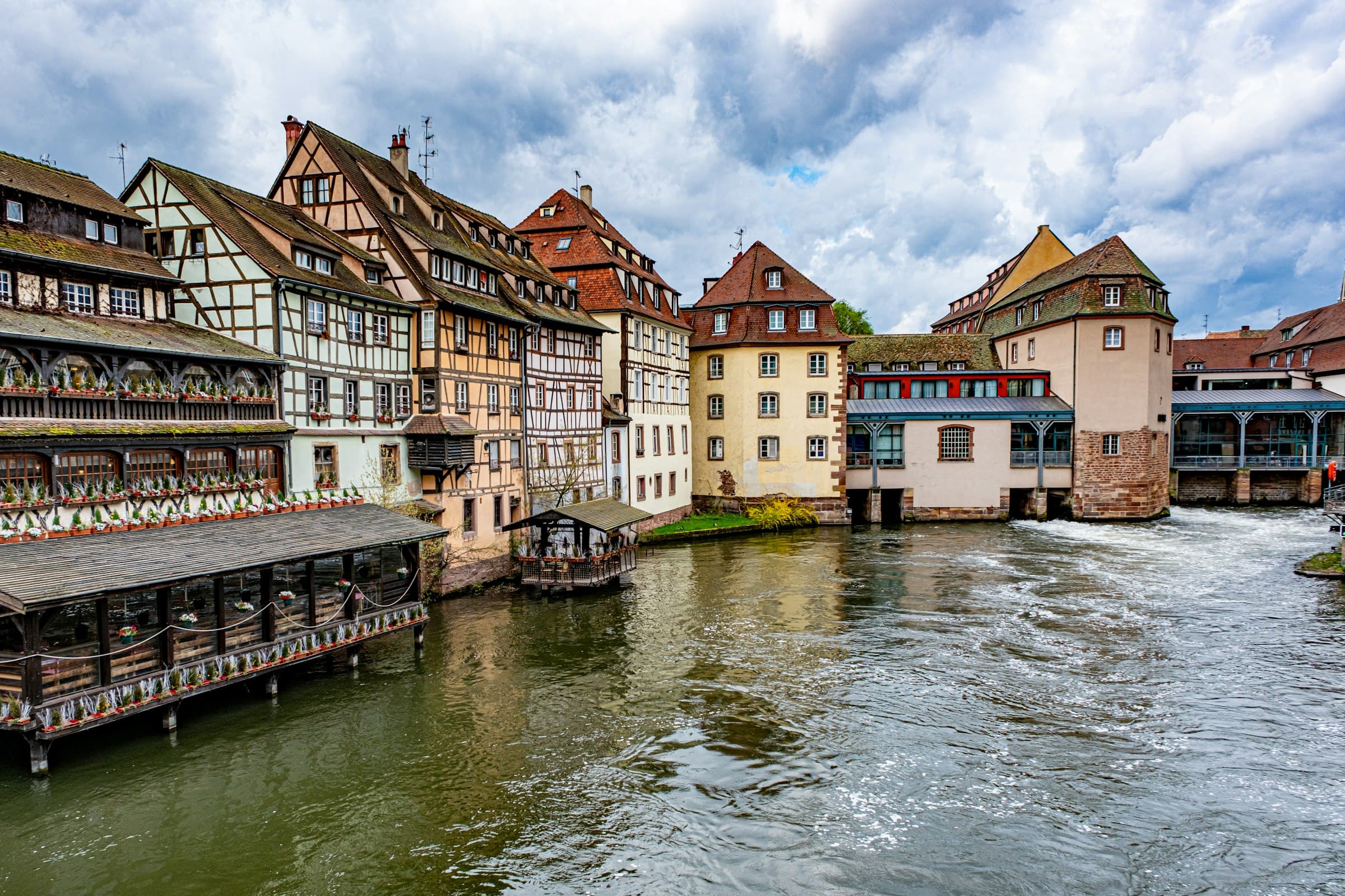 Un alignement de maisons à colombages de Strasbourg au bord de l’Ill, toits serrés et barrage au fil de l’eau sous un ciel nuageux